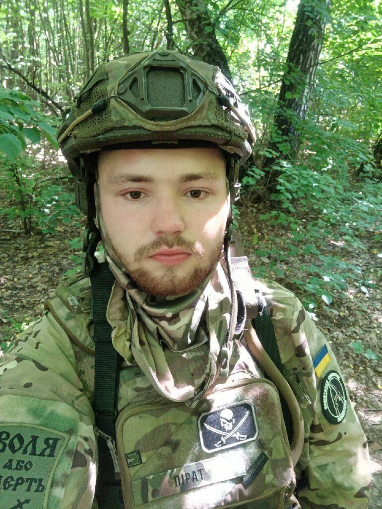  Vladislav "Pirate" Yurchenko in a wooded area wearing camo, with anarchist and ukranian patches, and a skull and cross swords 'jolly roger' patch on the chest. 