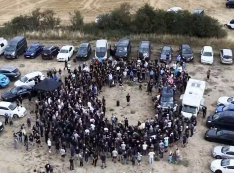 Thomas standing in a field surrounded by a circle of approximately 200 people, surrounded in turn by a couple of dozen vans and cars.